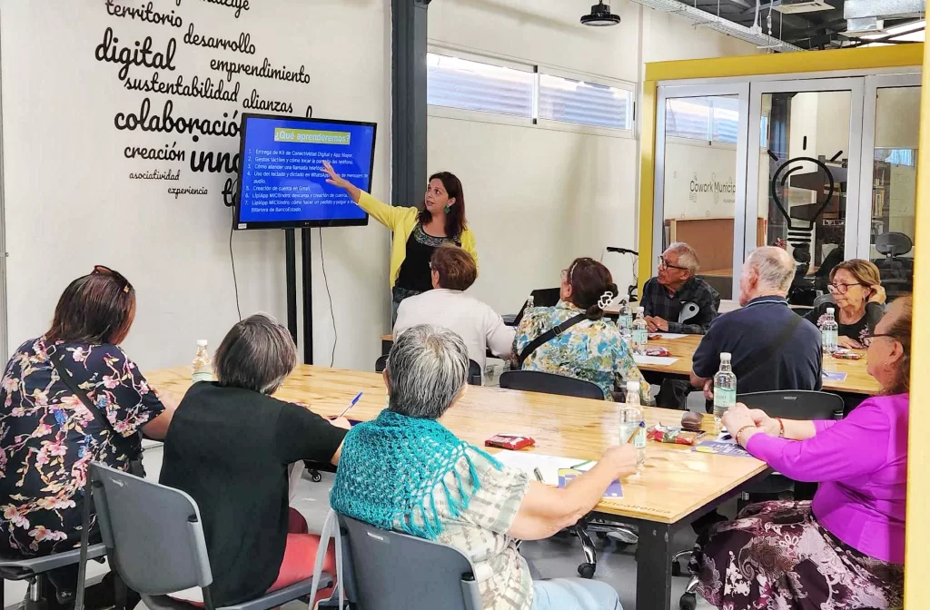 Una clase del Programa de Empoderamiento Digital, la profesora, que está rodeada de la menos 9 personas señala una pantalla de televisión con información teórica sobre cómo usar un teléfono inteligente. 