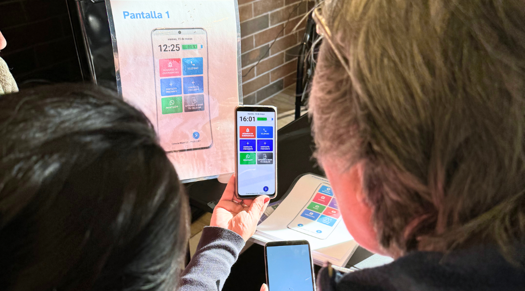 Fotografía muestra a dos personas desde la espalda mirando una lámina con la imagen de la pantalla 1 de App Mayor junto a un teléfono real con la misma pantalla de la aplicación.
